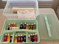 Wide shot of open mint green storage bin containing tubes of Artist Loft acrylic paint, with folded cream apron and roll of white paper placed inside a clear plastic container on the table.