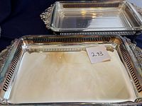 Two shiny silver-plated buffet trays, one stacked inside the other, with visible pierced gallery edge and ornate handles. The smaller tray contains a clear glass dish fitting inside it.