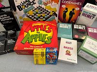 Photo showing a variety of games including Apples to Apples in red box, What Do You Meme? base game and expansion packs, Game Bounce party game, Codenames board game, and Cards Against Humanity boxes stacked on left.