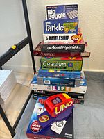 Stack of 13 board games showing games Big Boggle, Farkle, Battleship Grab & Go, Scattergories, Scrabble, Cranium, Connect 4, The Game of Life, Risk, UNO Attack, and Rummikub with visible box conditions.