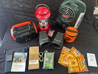 Full view of the camping supplies showing red Coleman lantern, black and orange portable power station, black hand warmers, orange carrying case, packets of hand warmers, solar accessory kit, user manuals, and small boxes.