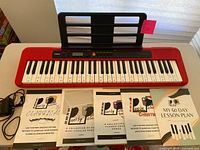 Red Casio Casiotone CT-S200 keyboard with music stand, power cord, and five piano lesson booklets arranged in front on a flat surface.