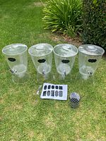 Four clear plastic drink dispensers with lids arranged on grass. Black adhesive label on each dispenser. Multiple shaped black labels and white marker placed in front of dispensers. Small black mesh container also visible.