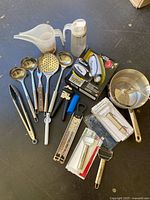 Assorted kitchen utensils including tongs, ladles, can openers, peelers, measuring jugs, and saucepan arranged on a dark surface.