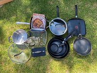 Cookware set laid out on grass showing frying pans, strainer, glass lids, baking dish, burger press