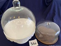 Two dome cover sets side by side on a dark blue cloth background. Left is a Decostar dome cover on a white metal cake stand. Right is a Hearth & Hand dome cover on a wood base.