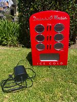 Front view of the red Drink-O-Magic mini fridge cooler outdoors on grass showing 6 circular window slots, 6 press buttons, and dispenser area.
