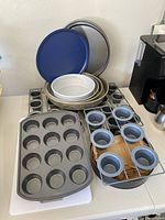 Variety of baking pans stacked including round cake pans, a springform pan, and large muffin pan with 12 muffin slots plus cooling racks and 2 grey mini muffin tins.