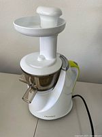Front view of the white Hurom H100 cold press juicer on a table showing its dual inlet hopper and juice spout.