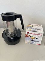 Electric coffee maker from Instant Pot with glass carafe, black plastic handle and base, beside box of 12-piece demitasse set.