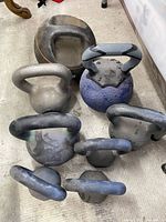 Eight kettlebells arranged on floor showing variety of sizes and styles from roughly 10 lb to 53 lb, clustered in group