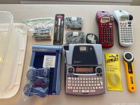 Full overview of the lot showing Brother P-Touch label maker, X-Acto rotary blade cutter with replacement blades, Dymo tape cartridges and parts.
