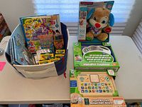 Full view of lot items arranged on table showing Leap Frog toys in box, Fisher Price Smart Stages Puppy in box, and a cloth bin filled with various Crayola coloring supplies and themed coloring books.