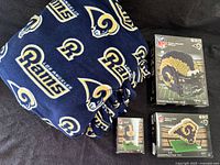 Photo showing folded navy blue Los Angeles Rams fleece blanket with gold and white logos beside three unopened BRXLZ NFL 3D construction toy boxes.