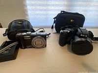 Photo showing Nikon Coolpix L120 camera with lens cover, Panasonic LUMIX DMC Z510 camera, chargers, cables, and carrying cases on a flat surface.