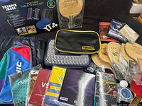 Photo showing multiple wooden table tennis paddles, some wrapped in plastic, various carrying cases, paddle covers, and DVDs arranged on a surface.