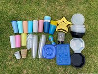Items spread on grass including plastic tall cups in pastel colors, clear disposable cups, smaller plastic cups, disposable food containers in round, star, hexagonal, and rectangular shapes, and plastic forks.