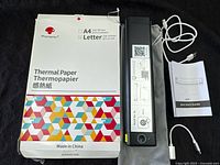 Photo of the black portable printer laying next to a large pack of A4 thermal paper sheets with multicolored geometric pattern on the packaging, along with a grey carrying pouch and white charging cable.