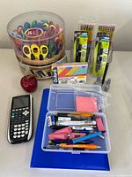Overview photo showing crafting scissors container, TI-84 calculator, packs of pencils, flashcards, tape dispenser, a red apple, and a small plastic box with assorted pens, erasers, and highlighter.