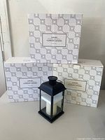 Three white boxes labeled 'Chelsea Solar Black Metal Flameless Lanterns Set of Two' with one black metal lantern with ivory flameless candle displayed in front.