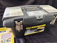 Closed Stanley 19" black plastic toolbox with label, two metal latches, handle, and clear plastic compartments on top for small parts.