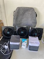 Five plug-in fans displayed alongside a grey fabric bin and a boxed light bulb on a surface.