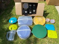 Lot of assorted plastic food storage containers, various sizes and shapes, mostly for cakes and cupcakes, arranged on grass with a cardboard box containing four black base containers visible.