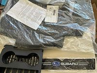 Black Subaru car floor mats in original plastic packaging with printed information sheet, black plastic dual cup holder insert, and Subaru logo sticker on carbon fiber patterned background.