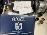 Photo showing a blue cover album titled 'The Official NFL Super Bowl Patch Collection' along with a framed display and part of several shot glasses.
