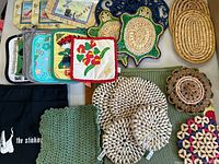 Top view showing assorted colorful pot holders, crocheted green holder, oval woven placemat, turtle-shaped woven trivets, and a set of printed fabric pot holders.