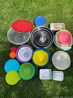 Photo shows various colorful plastic Tupperware containers with corresponding lids arranged on grass alongside a nested set of metal mixing bowls and foil containers.