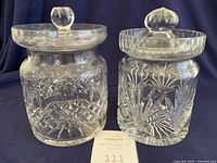 Two vintage crystal biscuit holders jars with lids, one Rogaska cut crystal and one resembling Waterford, shown side by side on dark cloth background.