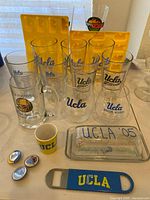 Lot of UCLA branded glassware, including pint glasses, football and basketball glasses, banquet mug, wine glasses, shot glass, Jello molds, butter dish, and bottle opener displayed on table.