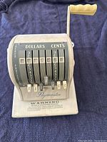Front view of a vintage Paymaster Series S-1000 check writer machine with beige/grey body. Visible dials labeled for dollars and cents and mechanical levers with metal tags showing currency symbols.