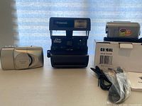 Wide shot of three cameras on table: Polaroid One Step, Olympus Stylus Epic Zoom 170, and Global DV 136ZB digital video camera with box and cables