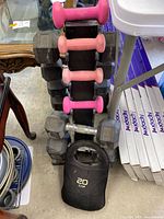 Photo showing black dumbbell rack with various dumbbells in pink neoprene and hexagonal rubber coated weights, and a 20 lb CAP kettlebell in a black cover on the floor.
