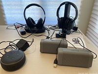 Lot of wireless headphones on charging stands, two portable AOMAIS Bluetooth speakers, and Amazon Echo Dot with cords on table