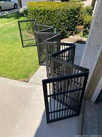 Full view of the folding gate laid out in an extended position on lawn and sidewalk, showing black vertical slat panels and multiple hinged sections.