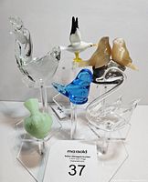 Seven vintage glass bird figurines arranged on acrylic display stands showing color, shape, and variety in the lot.