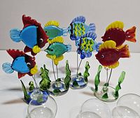 Four large colorful vintage art glass fish mounted on thin glass rods with round bases and green aquarium plant accents, positioned with floating glass bubbles nearby.