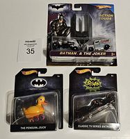 Photo of three Hot Wheels diecast cars in original Batman themed packaging, including Batman & The Joker Action Duos, The Penguin Duck, and Classic TV Series Batmobile.
