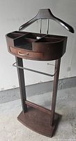 Front three-quarter view of the dark wooden bedroom valet stand showing hanger, storage compartments, drawer, and hanging bar.