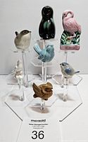 Front view of eight vintage bird figurines displayed on multiple transparent stands, showing variety of bird designs and colors