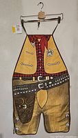 Front view of cowboy themed BBQ apron with yellow vest, red plaid shirt, sheriff badge, belt, and brown chaps design hung on hanger.