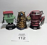 Front view of three Funko Pop robot figures: green and maroon Star Wars robot, bronze Doctor Who Dalek, red Star Wars robot with chains, all on display bases with Star Wars logos on two