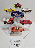 Front and angled view of all 7 Hot Wheels cars arranged on a clear display stand, showing variety in color and style.