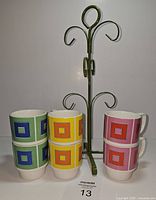 Six colorful stacking mugs arranged in three stacks by color beside a green display hanger on a white background.