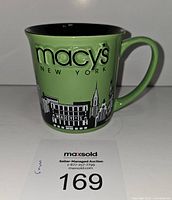 Side view of vintage green Macy's New York mug showing raised branding text, skyline buildings, and handle, with black interior.