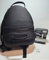 Front view of the black Polaroid-branded backpack with double zippers and adjustable straps, with Polaroid instant camera nearby.