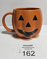 Front view of orange pumpkin-shaped mug with jack o' lantern face, showing handle and ceramic ridges.